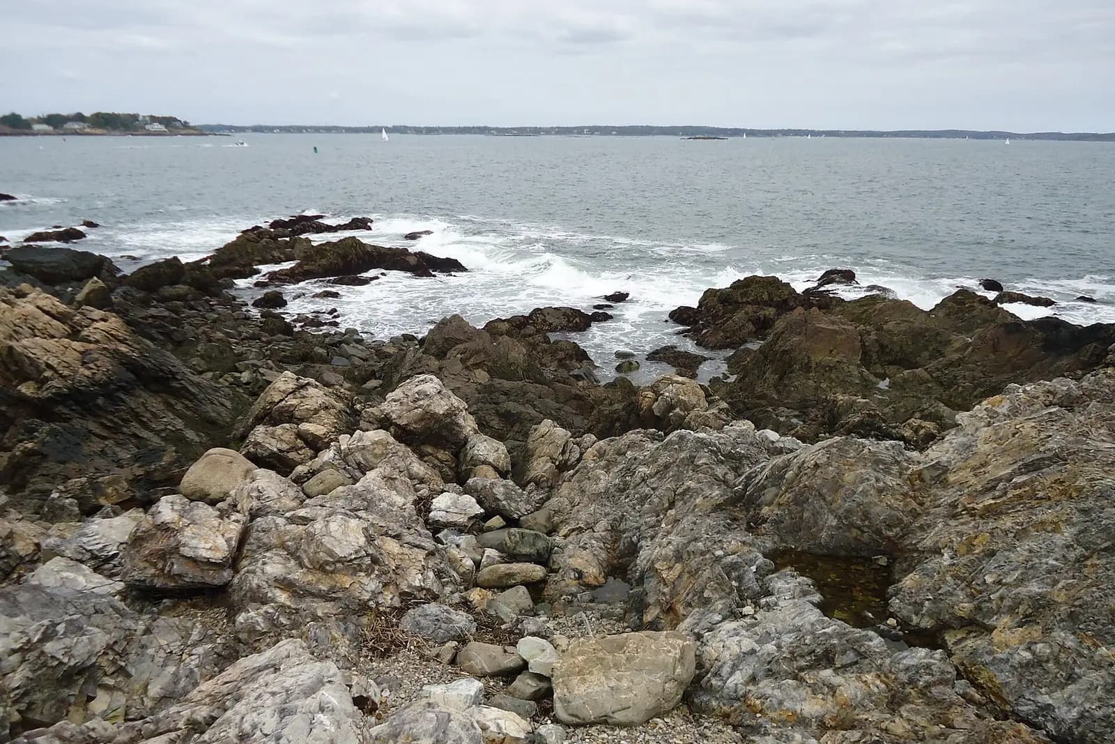 Granite rocks and breaking surf at the mouth of a harbour, with a wooded shoreline across the water.