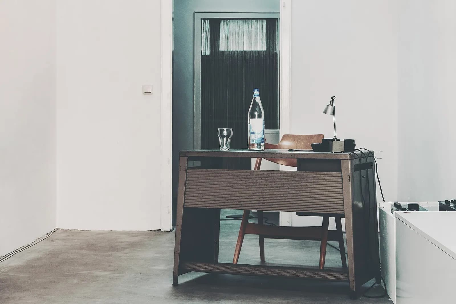 A wooden desk, a chair and a lamp in a plain, daylit room.